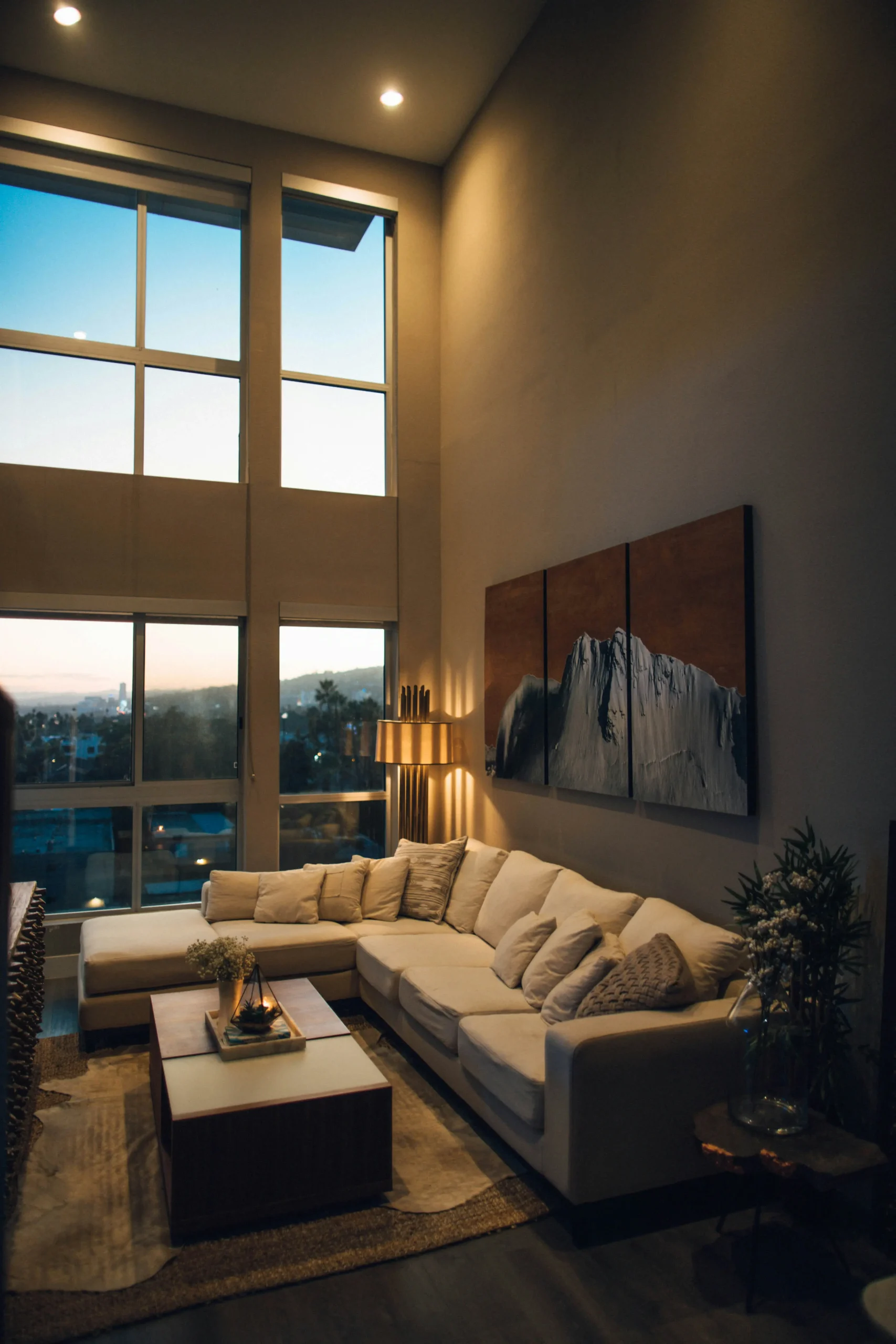 Salón a doble altura al atardecer, con sofá de lino y lámpara cálida junto a grandes ventanales