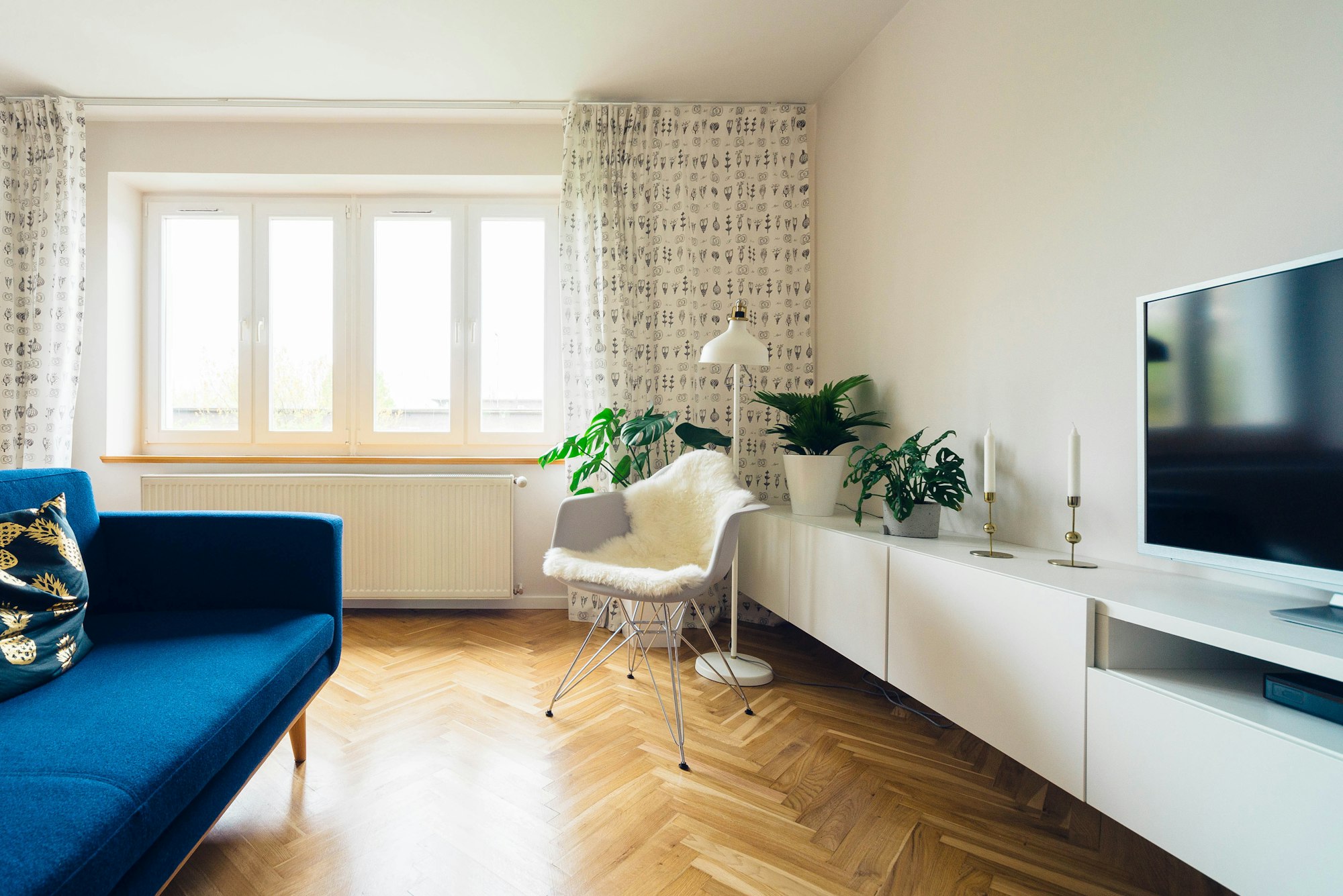 Salón con suelo de madera en espiga, sofá azul, mueble bajo blanco y una silla con piel de oveja junto a la ventana