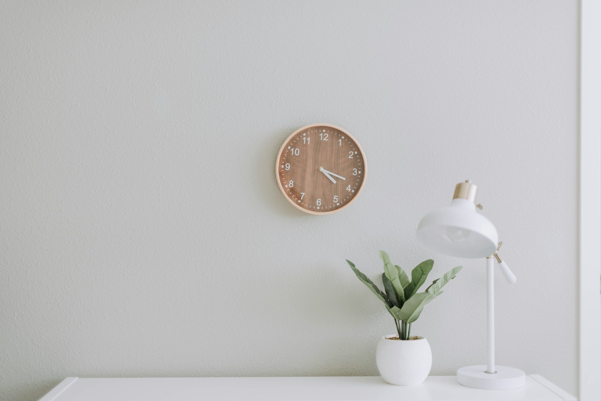 Escritorio blanco con lámpara de mesa, una planta y un reloj de madera en la pared